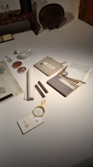 A collection of traditional writing materials is arranged on a tabletop, including scrolls, a brush and ink set, a magnifying glass, and books with Asian script. The setup suggests a historical or artistic context, possibly related to calligraphy or scholarly study.