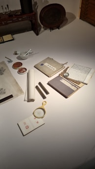 A collection of traditional writing materials is arranged on a tabletop, including scrolls, a brush and ink set, a magnifying glass, and books with Asian script. The setup suggests a historical or artistic context, possibly related to calligraphy or scholarly study.