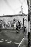 A group of young people are playing basketball on an outdoor court with a backdrop of a graffiti-covered wall. One player is jumping to shoot the basketball into the hoop, while others are standing around watching and participating. The scene captures movement and energy in a casual, urban setting.