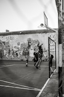 A group of young people are playing basketball on an outdoor court with a backdrop of a graffiti-covered wall. One player is jumping to shoot the basketball into the hoop, while others are standing around watching and participating. The scene captures movement and energy in a casual, urban setting.