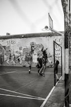 A group of young people are playing basketball on an outdoor court with a backdrop of a graffiti-covered wall. One player is jumping to shoot the basketball into the hoop, while others are standing around watching and participating. The scene captures movement and energy in a casual, urban setting.