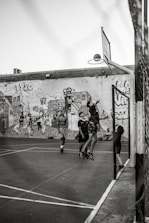A group of young people are playing basketball on an outdoor court with a backdrop of a graffiti-covered wall. One player is jumping to shoot the basketball into the hoop, while others are standing around watching and participating. The scene captures movement and energy in a casual, urban setting.