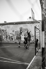 A group of young people are playing basketball on an outdoor court with a backdrop of a graffiti-covered wall. One player is jumping to shoot the basketball into the hoop, while others are standing around watching and participating. The scene captures movement and energy in a casual, urban setting.