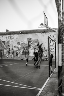 A group of young people are playing basketball on an outdoor court with a backdrop of a graffiti-covered wall. One player is jumping to shoot the basketball into the hoop, while others are standing around watching and participating. The scene captures movement and energy in a casual, urban setting.