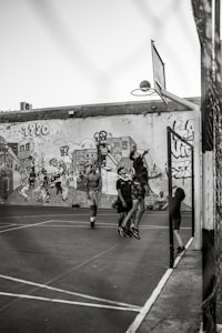 A group of young people are playing basketball on an outdoor court with a backdrop of a graffiti-covered wall. One player is jumping to shoot the basketball into the hoop, while others are standing around watching and participating. The scene captures movement and energy in a casual, urban setting.