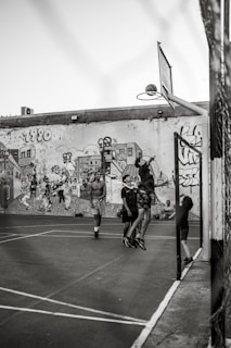 A group of young people are playing basketball on an outdoor court with a backdrop of a graffiti-covered wall. One player is jumping to shoot the basketball into the hoop, while others are standing around watching and participating. The scene captures movement and energy in a casual, urban setting.