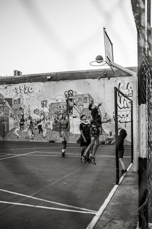 A group of young people are playing basketball on an outdoor court with a backdrop of a graffiti-covered wall. One player is jumping to shoot the basketball into the hoop, while others are standing around watching and participating. The scene captures movement and energy in a casual, urban setting.