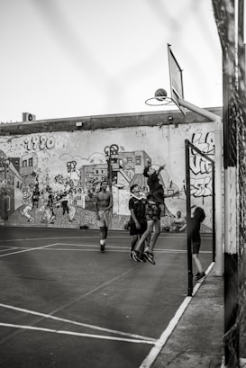 A group of young people are playing basketball on an outdoor court with a backdrop of a graffiti-covered wall. One player is jumping to shoot the basketball into the hoop, while others are standing around watching and participating. The scene captures movement and energy in a casual, urban setting.