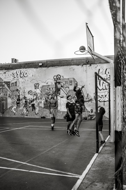A group of young people are playing basketball on an outdoor court with a backdrop of a graffiti-covered wall. One player is jumping to shoot the basketball into the hoop, while others are standing around watching and participating. The scene captures movement and energy in a casual, urban setting.