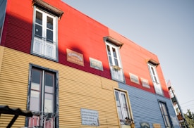 A brightly colored building features red, yellow, and blue corrugated panels. The structure includes several windows with white frames and small balconies. Signs with Spanish text are visible, adding a cultural element to the vibrant facade.