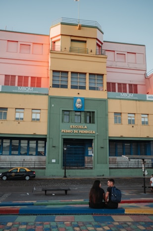 A colorful building with a central entrance labeled 'Escuela Pedro de Mendoza.' A couple is seated on a bench in the foreground on a multicolored pavement. The facade of the building features various pastel hues, including yellow, green, and pink.