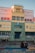 A colorful building with a central entrance labeled 'Escuela Pedro de Mendoza.' A couple is seated on a bench in the foreground on a multicolored pavement. The facade of the building features various pastel hues, including yellow, green, and pink.