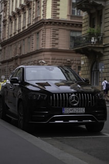 A black luxury car parked on the street in front of an ornate, historic building. The car is sleek and modern, with a visible license plate and distinct grille design. The architecture of the building is classic with detailed cornices and tall, narrow windows.