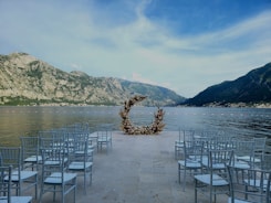 An outdoor wedding setup is situated by a lake surrounded by steep mountains. A circular floral arch forms a focal point at the end of an aisle lined with rows of white chairs.