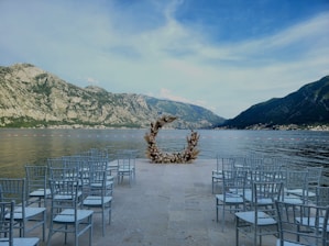 An outdoor wedding setup is situated by a lake surrounded by steep mountains. A circular floral arch forms a focal point at the end of an aisle lined with rows of white chairs.