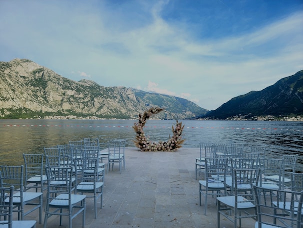 An outdoor wedding setup is situated by a lake surrounded by steep mountains. A circular floral arch forms a focal point at the end of an aisle lined with rows of white chairs.