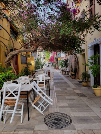 A charming narrow alley with outdoor seating arrangements featuring white wooden chairs and tables. The alley is adorned with vibrant bougainvillaea plants hanging over the pathway and potted plants along the sides. It is surrounded by warm-toned buildings with windows and shutters.