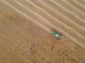 A green agricultural combine harvester is cutting a swath through a large, golden wheat field, leaving behind harvested rows. The aerial perspective emphasizes the vastness of the farmland and the machine's precision as it moves through the crops.