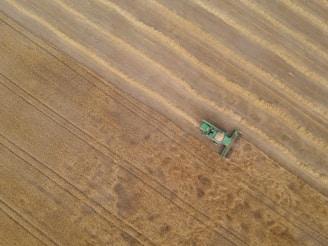 A green agricultural combine harvester is cutting a swath through a large, golden wheat field, leaving behind harvested rows. The aerial perspective emphasizes the vastness of the farmland and the machine's precision as it moves through the crops.