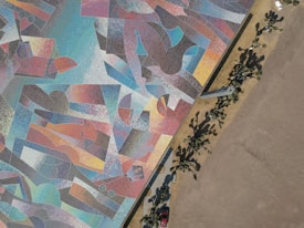 Aerial view of a large, colorful mosaic artwork featuring abstract geometric patterns and shapes. The mosaic covers a significant portion of the ground next to a barren area. On the right, there are shadows of palm trees cast on a paved walkway, with a red car parked nearby.