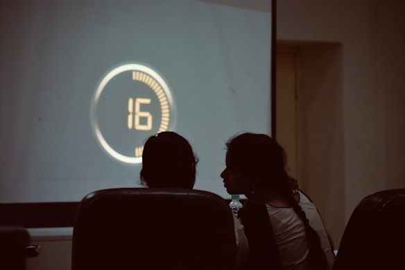 Two people are seated in a dimly lit room, facing a large screen displaying a countdown timer with the number 16 in a circular design.
