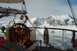 A vintage wooden ship is navigating through icy waters with a snowy mountain range in the background. The ship's deck features a large steering wheel, sails, and ropes, suggesting a classic maritime setting.