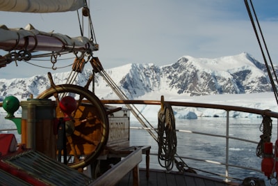 A vintage wooden ship is navigating through icy waters with a snowy mountain range in the background. The ship's deck features a large steering wheel, sails, and ropes, suggesting a classic maritime setting.