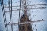 A tall ship navigates through icy waters with sails down against a backdrop of an expansive ice field. The rigging and masts are visible, with several crew members working aboard the deck, surrounded by a cold, arctic environment.