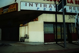 A street scene featuring an exterior view of a building with signage for a Peruvian restaurant. The facade includes a partially visible open sign, metal gates, and a graffiti mark on the wall. There are shadows cast on the building, suggesting strong sunlight. A section of the sidewalk and a utility pole are also visible.