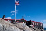 A group of penguins are perched on rocky terrain in front of a black and red building with a Union Jack flag flying above. The sky is clear and blue with a few scattered clouds. The building has several windows with white frames and a staircase leading up to an entrance.
