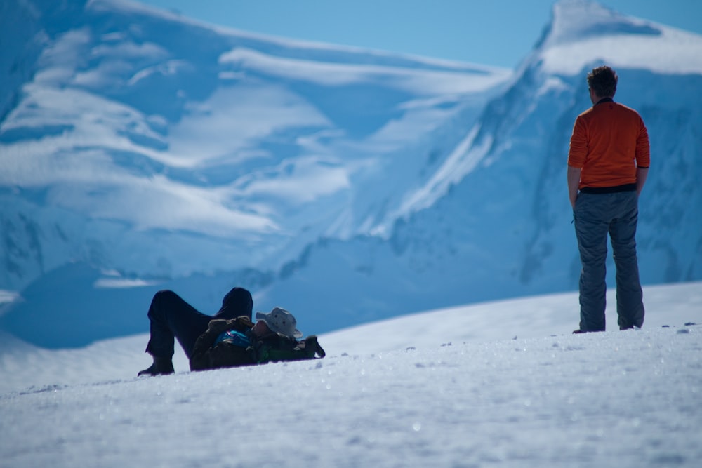 a man standing next to a woman laying on top of a snow covered slope