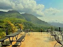 A spacious terrace with outdoor seating, including tables, chairs, and picnic benches, overlooks lush green mountains under a partly cloudy sky. Various potted plants add to the serene atmosphere, enhancing the connection with the natural surroundings.