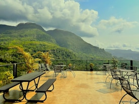 A spacious terrace with outdoor seating, including tables, chairs, and picnic benches, overlooks lush green mountains under a partly cloudy sky. Various potted plants add to the serene atmosphere, enhancing the connection with the natural surroundings.