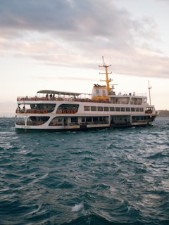A large ferry navigates through choppy waters with a partly cloudy sky overhead. The vessel is adorned with numerous life rings along its sides, and there are people visible on the upper deck. The ferry has a distinctive yellow and white color scheme, with a prominent yellow smokestack. The scene conveys a sense of movement as the ship travels across the sea.
