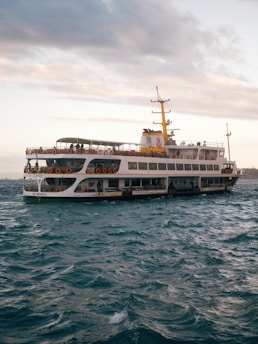 A large ferry navigates through choppy waters with a partly cloudy sky overhead. The vessel is adorned with numerous life rings along its sides, and there are people visible on the upper deck. The ferry has a distinctive yellow and white color scheme, with a prominent yellow smokestack. The scene conveys a sense of movement as the ship travels across the sea.