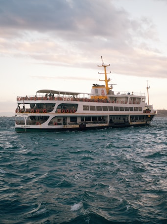 A large ferry navigates through choppy waters with a partly cloudy sky overhead. The vessel is adorned with numerous life rings along its sides, and there are people visible on the upper deck. The ferry has a distinctive yellow and white color scheme, with a prominent yellow smokestack. The scene conveys a sense of movement as the ship travels across the sea.