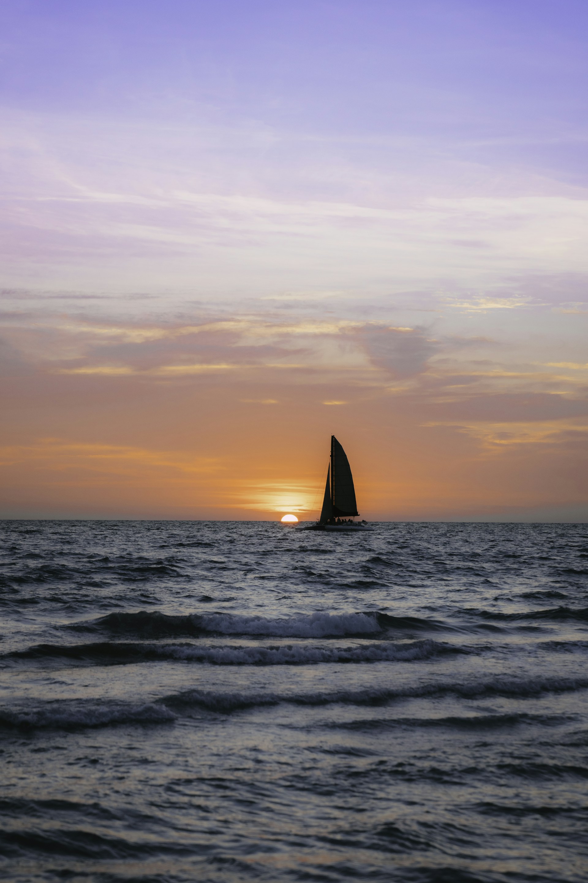 The silhouette of a sailboat framed by the glowing colors of a fading sunset, with soft waves around.