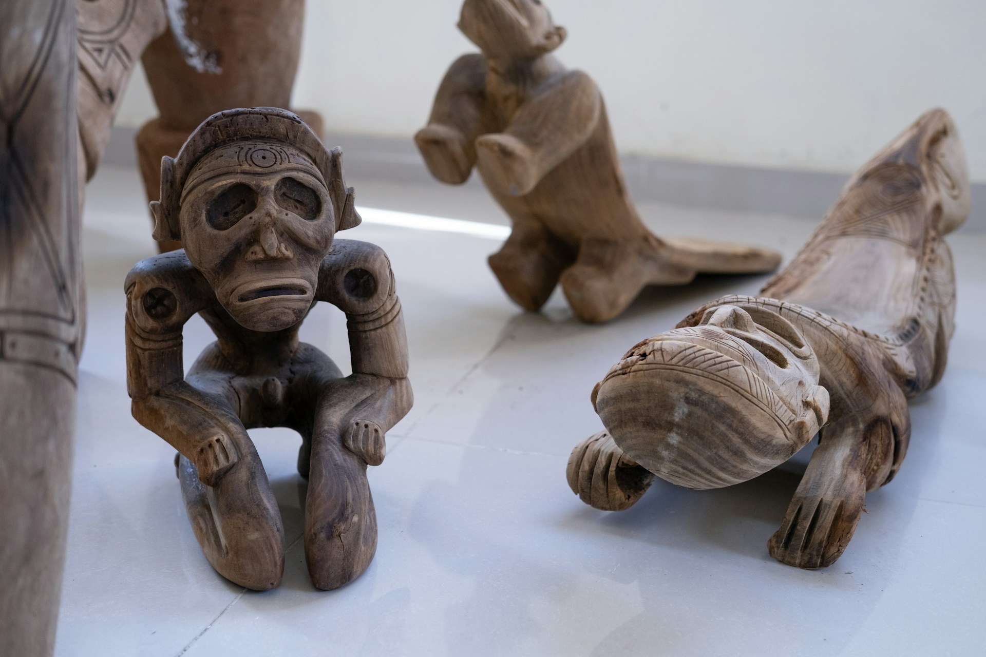 A collection of intricately carved wooden sculptures featuring humanoid figures. The central figure is kneeling, with a solemn expression and detailed carvings on its arms and head. Another figure is lying down, showcasing similar detailed craftsmanship. The sculptures are placed on a smooth surface with soft lighting accentuating their features.