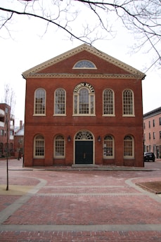 A historic brick building labeled 'Town Hall' with tall arched windows and a large central door, surrounded by a cobblestone plaza and leafless tree branches against a cloudy sky.