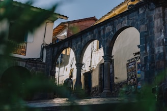 An old stone archway stands prominently with three arches, set against a backdrop of traditional buildings with tiled roofs. The area seems to be a historical or cultural site with signs and posters visible on the wall. Some greenery is present in the foreground, slightly out of focus.