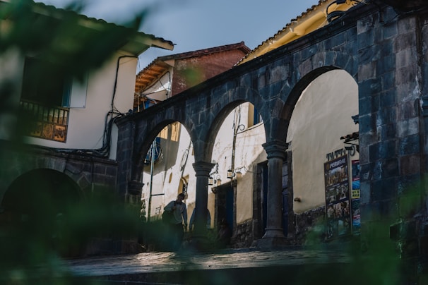 An old stone archway stands prominently with three arches, set against a backdrop of traditional buildings with tiled roofs. The area seems to be a historical or cultural site with signs and posters visible on the wall. Some greenery is present in the foreground, slightly out of focus.