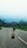 A person is walking along a deserted road, carrying a suitcase and wearing a backpack. The road is surrounded by lush green trees and the sky is a clear blue. The scene evokes a sense of solitude and adventure as the person heads towards an unknown destination.