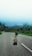 A person is walking along a deserted road, carrying a suitcase and wearing a backpack. The road is surrounded by lush green trees and the sky is a clear blue. The scene evokes a sense of solitude and adventure as the person heads towards an unknown destination.