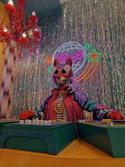 A figure dressed as a DJ stands behind a mixing table in a vibrant space with a disco theme. The figure wears eccentric clothing, styled with sunglasses and a colorful wig. Neon signs and shimmering metallic curtains create a festive and retro atmosphere. A bright red chandelier and a mirrored disco ball add to the decor.
