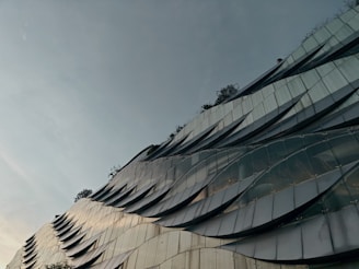 A modern building facade featuring a wavy, metallic design with reflective panels that create a dynamic visual texture. Sparse greenery is visible at the top, adding a natural element to the architectural structure. The sky is overcast, contributing to a subdued atmosphere.