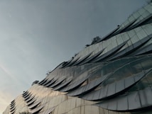 A modern building facade featuring a wavy, metallic design with reflective panels that create a dynamic visual texture. Sparse greenery is visible at the top, adding a natural element to the architectural structure. The sky is overcast, contributing to a subdued atmosphere.