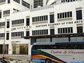 The image displays a large building with the signage 'Nanyang Academy of Fine Arts'. The structure is modern with multiple windows and the sign prominently displayed at the top. The ground level features a section labeled as NAFA Campus 2 with clear glass doors and an entrance. In the foreground, a bus with colorful stripes and the words 'Explore & Discover' on its side is present.