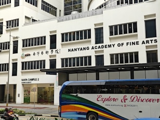 The image displays a large building with the signage 'Nanyang Academy of Fine Arts'. The structure is modern with multiple windows and the sign prominently displayed at the top. The ground level features a section labeled as NAFA Campus 2 with clear glass doors and an entrance. In the foreground, a bus with colorful stripes and the words 'Explore & Discover' on its side is present.