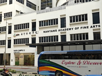 The image displays a large building with the signage 'Nanyang Academy of Fine Arts'. The structure is modern with multiple windows and the sign prominently displayed at the top. The ground level features a section labeled as NAFA Campus 2 with clear glass doors and an entrance. In the foreground, a bus with colorful stripes and the words 'Explore & Discover' on its side is present.