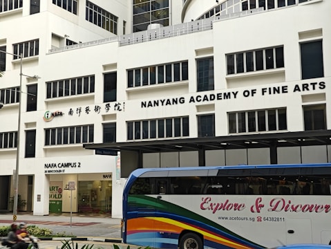 The image displays a large building with the signage 'Nanyang Academy of Fine Arts'. The structure is modern with multiple windows and the sign prominently displayed at the top. The ground level features a section labeled as NAFA Campus 2 with clear glass doors and an entrance. In the foreground, a bus with colorful stripes and the words 'Explore & Discover' on its side is present.